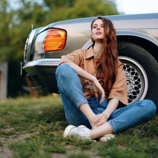 A stylish young woman with long hair sits beside a vintage car, dressed in casual attire. She reflects thoughtfully against a serene outdoor backdrop, embodying a relaxed and nostalgic vibe.